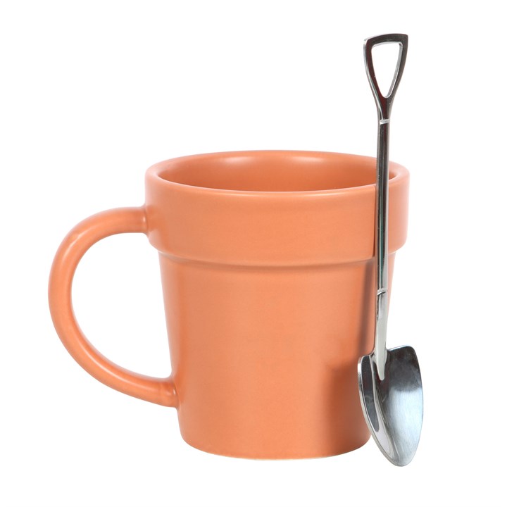 Terracotta orange ceramic mug shaped like a plant pot on a wooden table, with a shiny stainless steel mini shovel spoon resting against the rim on a white background