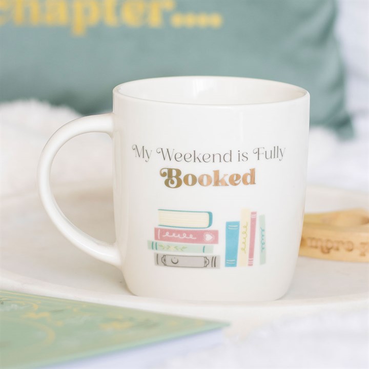 White ceramic mug with the phrase 'My Weekend is Fully Booked' and a pastel illustration of books, sitting on a white surface with a blurred green background.