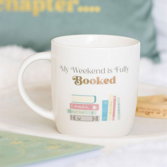 White ceramic mug with the phrase 'My Weekend is Fully Booked' and a pastel illustration of books, sitting on a white surface with a blurred green background.