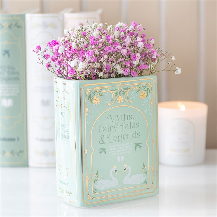 Sage green ceramic vase shaped like an upright fairytale book reading 'Myths, Fairy Tales, & Legends', holding vibrant pink and white flowers on a desk.