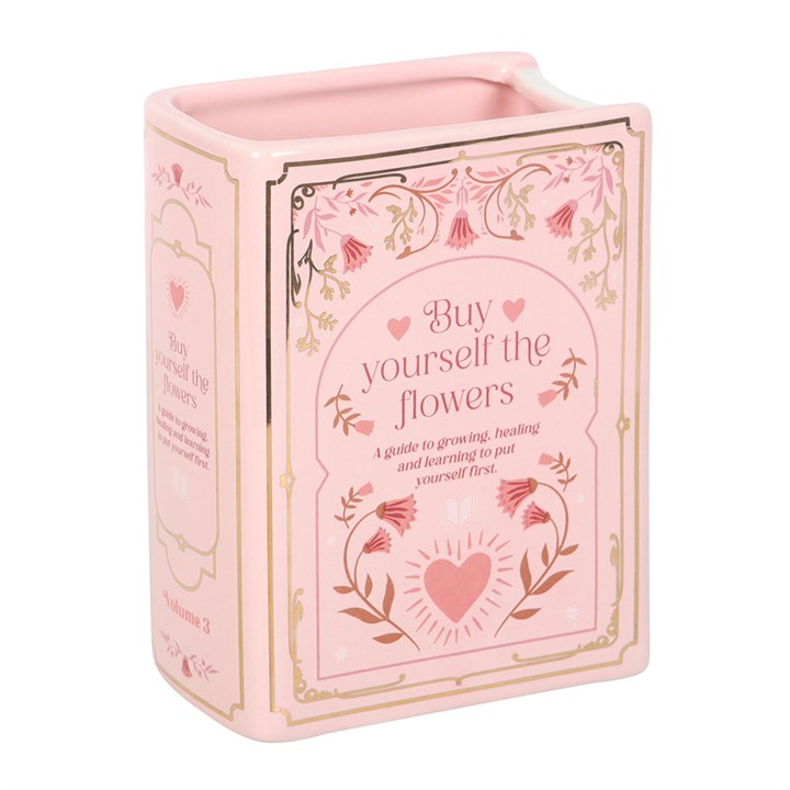 Pastel pink ceramic vase shaped like an upright book with  illustrations of a heart and text 'Buy Yourself the Flowers' on white background.