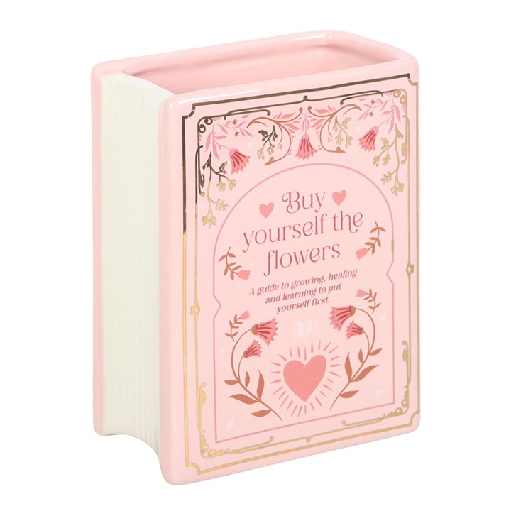 Pastel pink ceramic vase shaped like an upright book with  illustrations of a heart and text 'Buy Yourself  the Flowers' showing white pages end of the vase on white background.