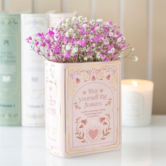 Pastel pink ceramic vase shaped like an upright book with illustrations of a heart and floral details, holding pink and white baby's breath flowers on a white surface