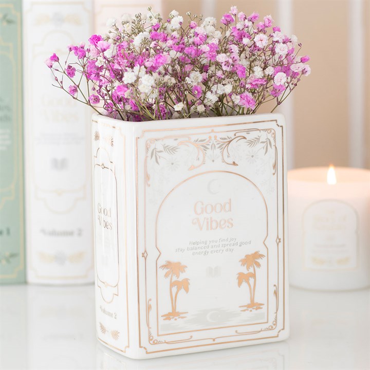 White ceramic vase shaped like an upright book with gold illustrations of palm trees and 'Good Vibes Volume 2' printed on the spine.