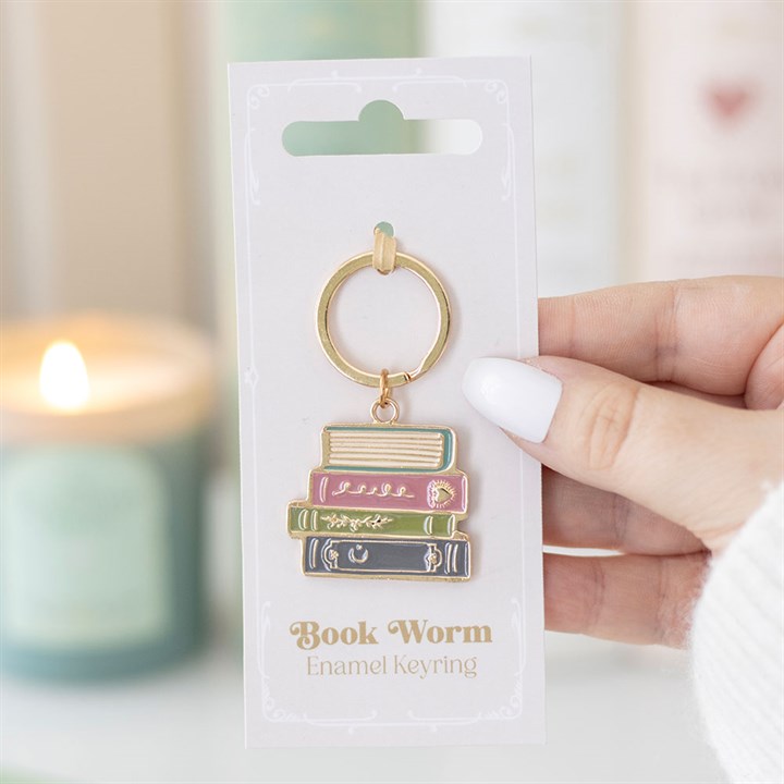 Close-up of a hand holding the 'Book Worm' enamel keyring, showing a pastel stack of four books on a white gift backing card, with a blurred blue candle in the background.