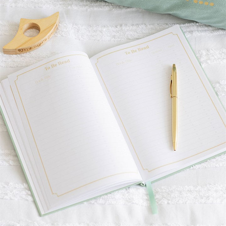 Open hardcover reading journal showing structured 'To Be Read' tracking log list pages with gold borders and a mint green ribbon page marker.