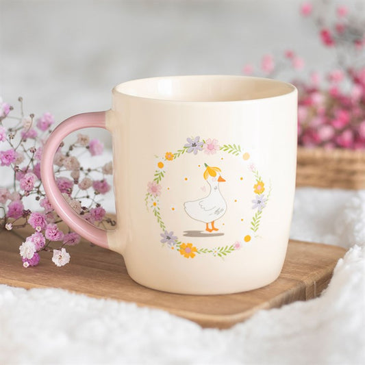Soft cream ceramic mug featuring a white duck inside a colorful wildflower wreath with a pastel pink handle, sitting on a rustic wooden board next to pink flowers.
