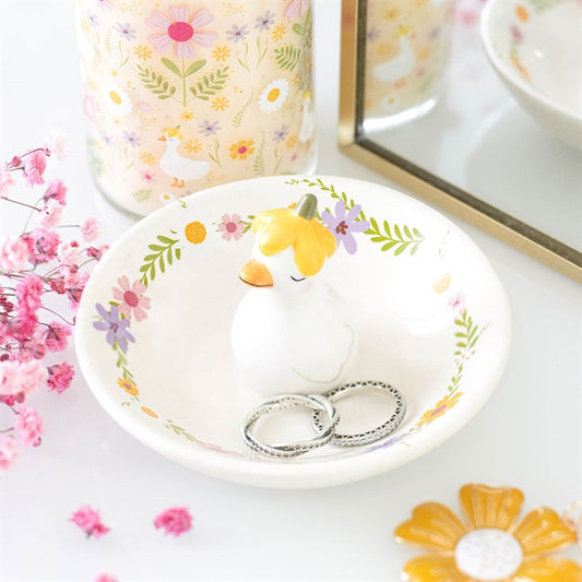 A small white ceramic trinket dish with a pastel wildflower border and a 3D duck figure in a yellow flower hat in the center, holding two silver rings on a vanity table.