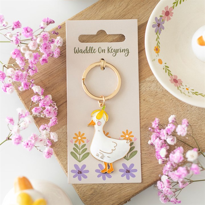 White enamel duck keyring wearing a yellow flower hat, mounted on a cream gift backing card with pastel illustrations, resting on a wooden board next to pink flowers.