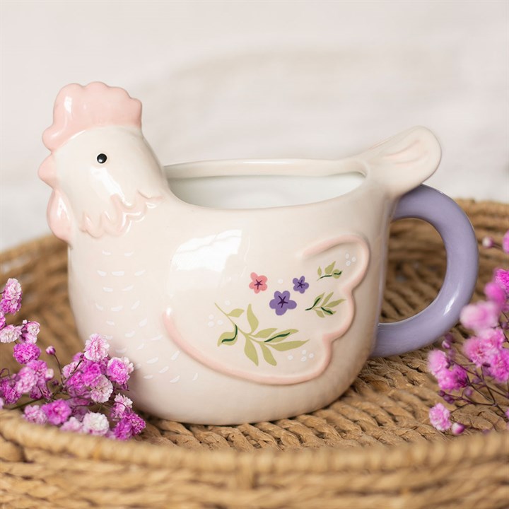 A sculpted cream ceramic mug shaped like a hen with a pink comb and lilac handle, featuring pastel floral paintings on its wings, sitting on a woven wicker tray next to pink flowers.