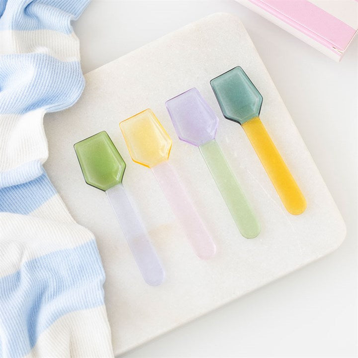 Four translucent glass dessert spoons in pastel green, yellow, lilac, and blue, arranged on a white marble board next to a striped blue cloth.