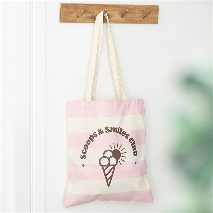 Pink and cream horizontal striped tote bag with 'Scoops & Smiles Club' ice cream logo on the front, hanging on a wooden peg.