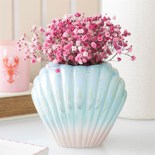 Pearlescent blue and pink ceramic shell vase holding pink flowers, sitting on a white table next to a lobster candle.