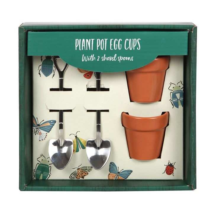 A green botanical gift display box containing two terracotta orange ceramic egg cups shaped like plant pots and two stainless steel mini shovel spoons on a white background.