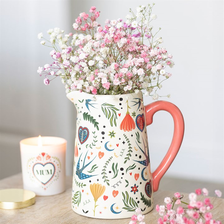 Cream ceramic jug with a red handle, covered in a colorful folk art print of wildflowers and birds, holding pink gypsophila stems on a rustic table.