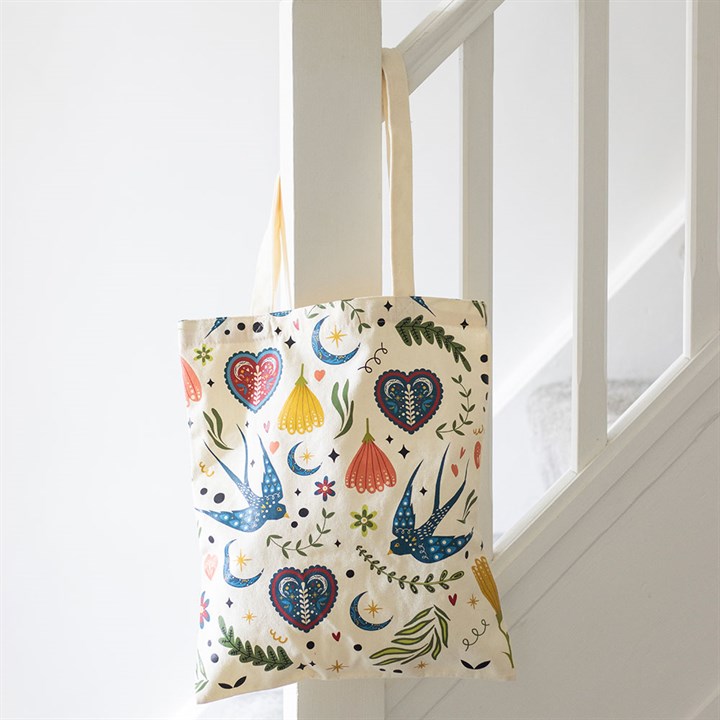 Cream canvas polycotton tote bag covered in a colorful folk art print of blue swallows, flowers and hearts, hanging from a white wooden staircase banister.