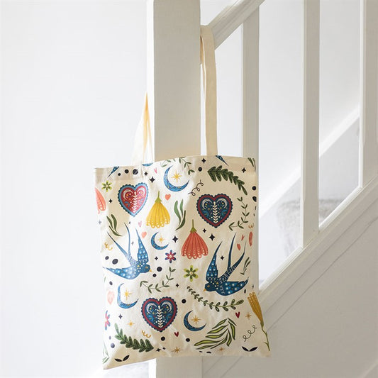 Cream canvas polycotton tote bag covered in a colorful folk art print of blue swallows, flowers and hearts, hanging from a white wooden staircase banister.