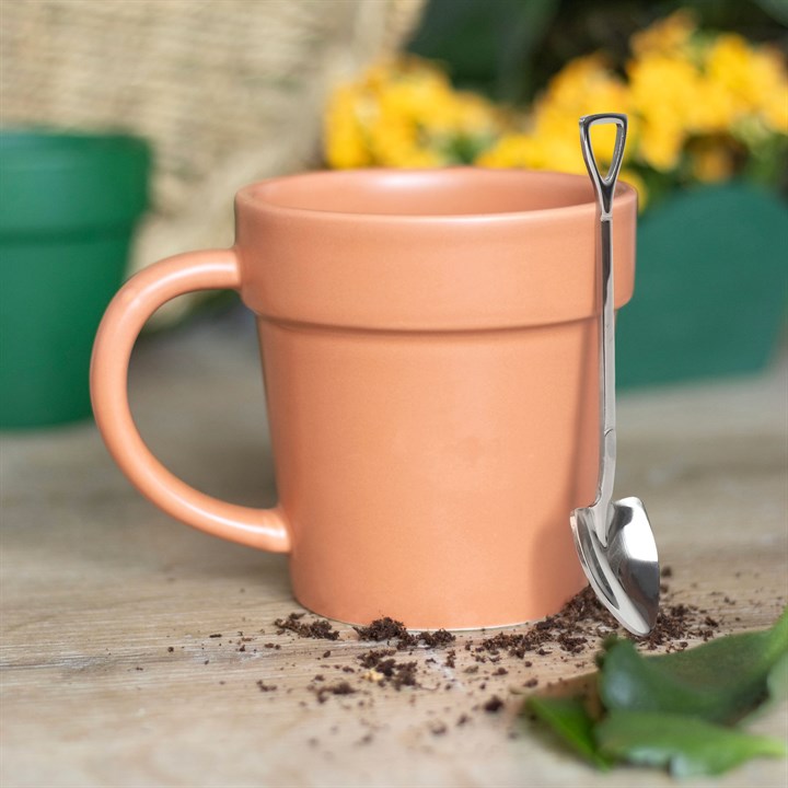 Terracotta orange ceramic mug shaped like a plant pot on a wooden table, with a shiny stainless steel mini shovel spoon resting against the rim.