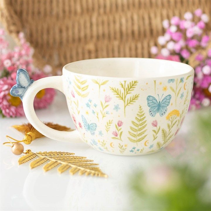 A wide white ceramic mug with a full-wrap print of pastel wildflowers and ferns, featuring a small 3D blue ceramic butterfly sculpted onto the handle, resting on a white surface.